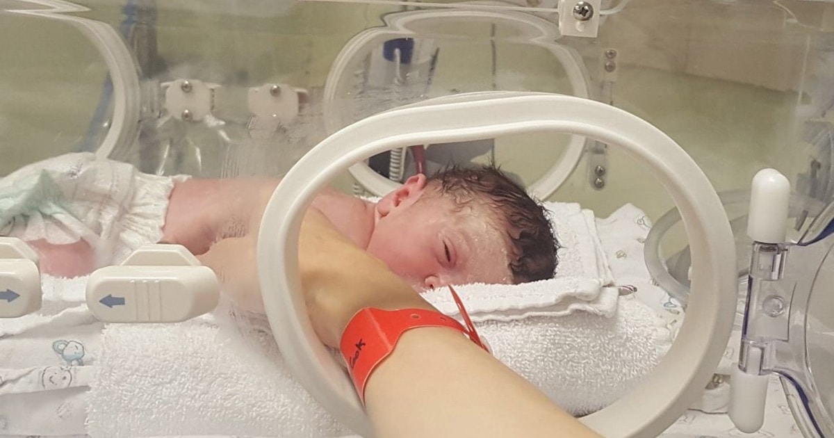 A baby in a premature baby incubator, with parent's hand reaching into porthole.