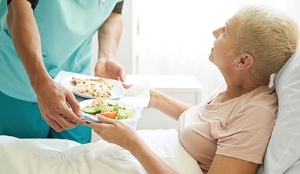 A hospital worker hands a tray of food to a patient sitting in bed.